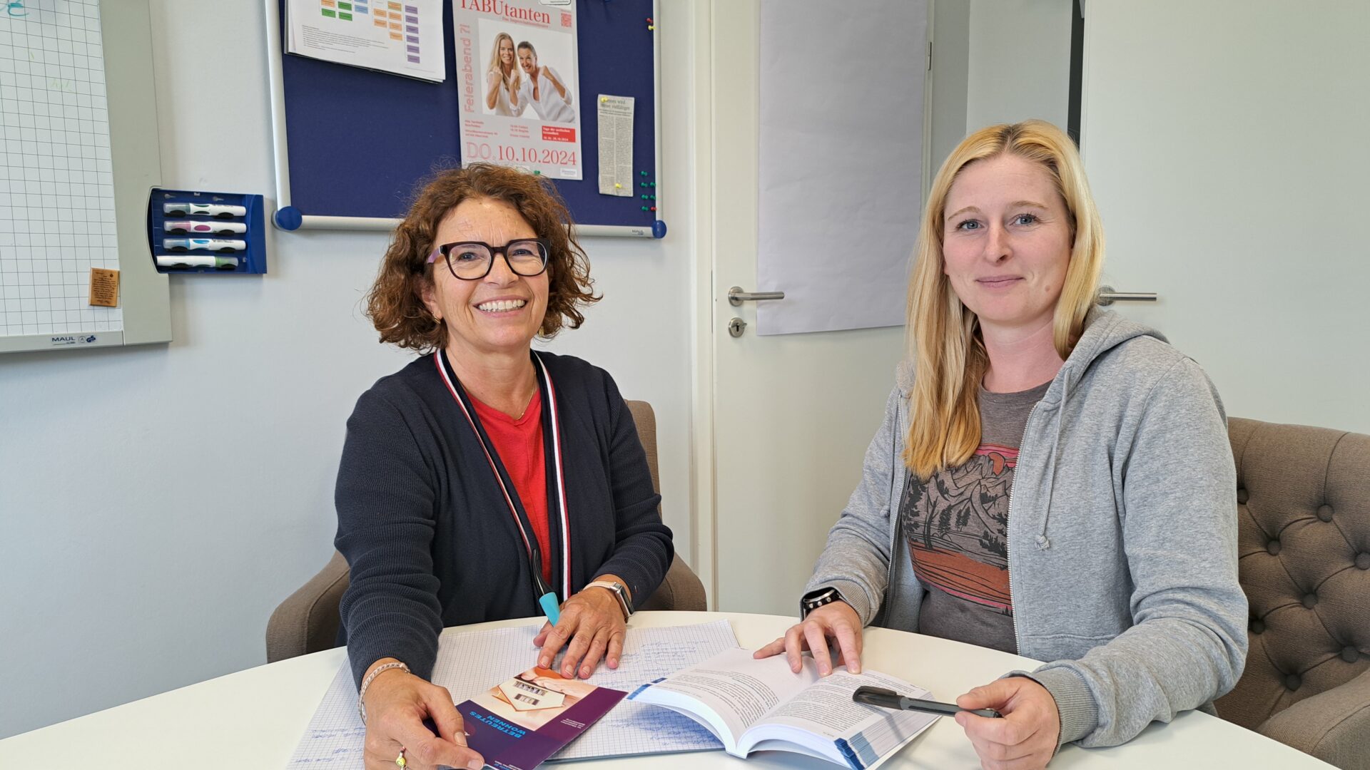 Zwei Frauen sitzen gemeinsam an einem Tisch und lachen in die Kamera.