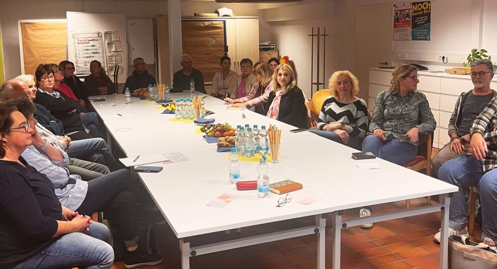 Mehrere Personen sitzen an einer langen Tafel und hören einem Vortrag zu.